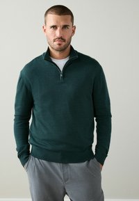 Mörkgrön zip-up-tröja i mjukt tyg med hög krage och ribbad fåll, bärs över en vit skjorta och kombineras med grå byxor.