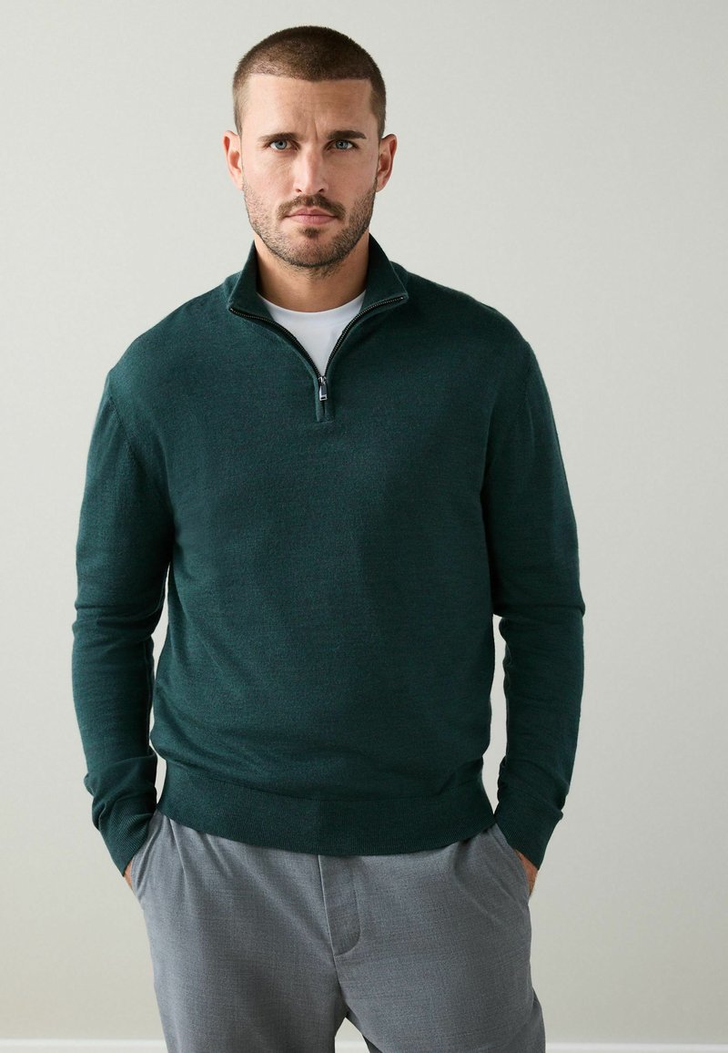 Mörkgrön zip-up-tröja i mjukt tyg med hög krage och ribbad fåll, bärs över en vit skjorta och kombineras med grå byxor.