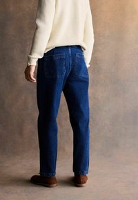 Jeans de mezclilla azul con un corte ajustado, que presentan dos bolsillos traseros y un diseño limpio. Se muestra con un suéter color crema y zapatos marrones.