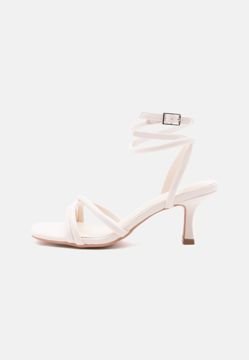 Hvid sandal med hæl, spænde-front, ankelrem med spænde og en slank mellemhøj hæl mod en ensfarvet baggrund.