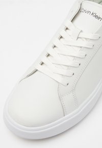 Sneaker bianco in pelle con lacci piatti, superficie liscia, punta arrotondata e etichetta con marchio. Collo imbottito e suola testurizzata per una migliore aderenza.