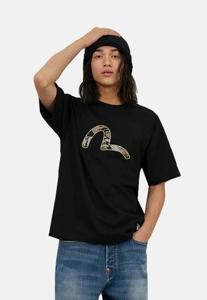 Sort bomulds T-shirt med et trykt abstrakt design i guld og brune nuancer. Korte ærmer og en løs pasform. Denimbukser bæres sammen med skjorten.