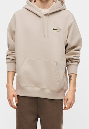 Mann trägt beigen Nike Hoodie mit Fronttasche und braunen Hosen, mit sichtbarem Tattoo am linken Unterarm.