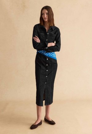 Vrouw draagt een lange zwarte denimjurk met knopen en een blauw-wit geruite sjaal gestrikt rond de taille, staand met gekruiste armen.