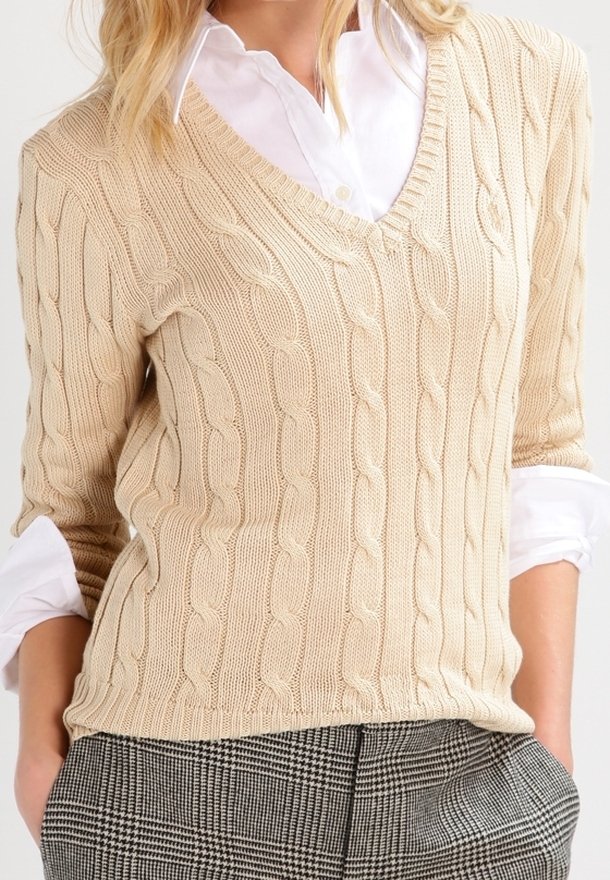 Pull beige en maille torsadée à col en V porté sur une chemise blanche à col, associé à un pantalon à carreaux noir et blanc.