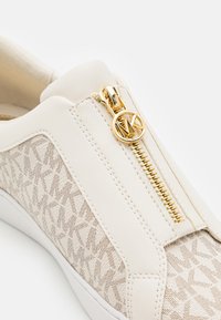 Baskets couleur crème avec motif texturé du logo MK, fermeture éclair dorée et tirette en forme de logo MK rond sur le côté.