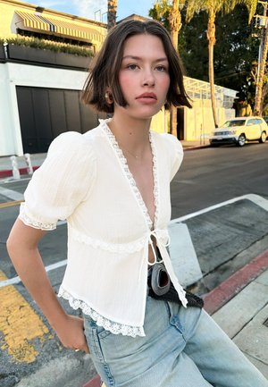 Tânără cu păr scurt și castaniu, purtând o bluză albă cu dantelă și blugi albaștri deschiși, stă pe marginea trotuarului într-o zi luminoasă.