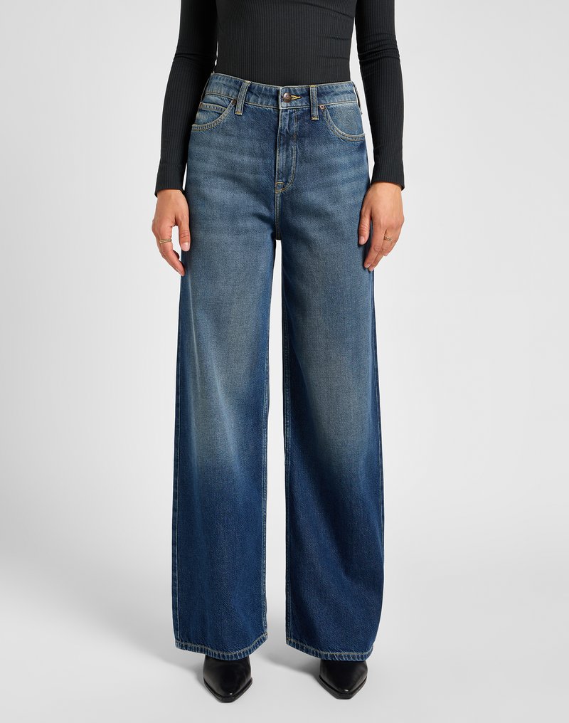 Woman wearing high-waisted wide-leg blue jeans, black ribbed long-sleeve top, black pointed shoes, standing against a plain light background.
