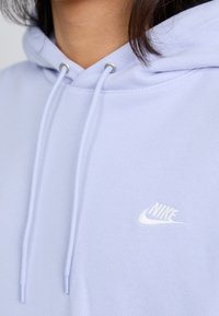 A close-up of a person wearing a light purple hoodie, featuring a white Nike logo and drawstrings on the hood. Hair is partially visible.