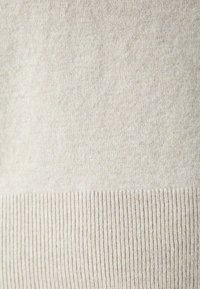 Beige gestrickter Pulloverstoff mit einer glatten Textur und geripptem Saum. Das Material wirkt warm und leicht, mit subtilen Farbabstufungen.