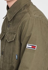 Olive green cotton jacket featuring a button-up design, two chest pockets, and a branded patch with red, white, and blue stripes on the sleeve.