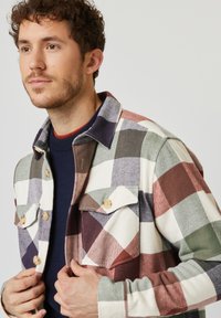 Geruite jas in crème, groen, bordeauxrood en marineblauw met twee borstzakken, ronde kraag en houten knopen; zachte textuur, ruime pasvorm.