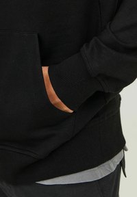 Sweat-shirt noir avec des poignets et un ourlet côtelés ; doté d'une poche avant. Tissu en coton texturé visible, superposé à une chemise grise.