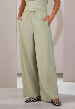 Person trägt hellgrüne, weit geschnittene Hose mit Kordelzug in der Taille, Hände in den Taschen und schwarz-weiß gemusterte Ballerinas.