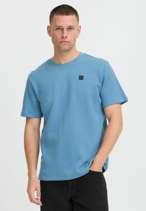 Young man with short hair wears a textured light blue short-sleeve shirt and black pants, standing with one hand in pocket.