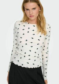 Biała bluzka z długim rękawem o żebrowej fakturze, z rozproszonym kwiatowym wzorem w odcieniach granatowego i zielonego oraz falbaniastym wykończeniem dołu.