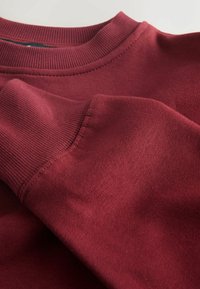 Burgunderfärgad sweatshirt med ribbad rundad hals och muddar. Mjuk textur, slätt tyg och synliga sömnadsdetaljer på ärmarna.