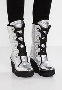 Stříbrné kotníkové wedge boty po kolena s černými šňůrkami a lemy, nošené s černými legínami, s viditelným logem Calvin Klein na jazycích.