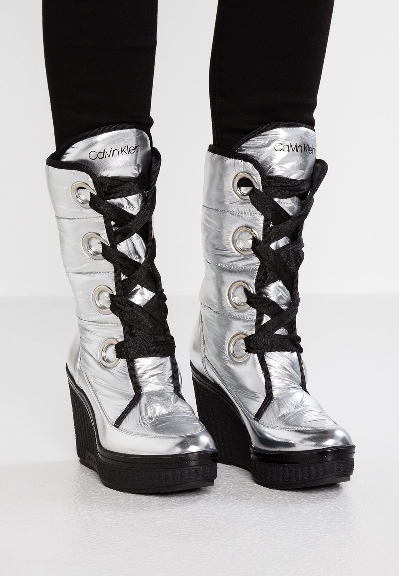Stříbrné kotníkové wedge boty po kolena s černými šňůrkami a lemy, nošené s černými legínami, s viditelným logem Calvin Klein na jazycích.