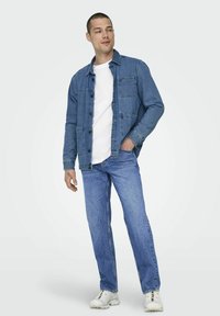 Chaqueta de denim en azul con cierre de botones, dos bolsillos en el pecho y un corte casual, combinada con jeans de azul claro y zapatillas blancas.