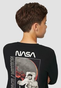 Persoon met kort bruin haar, gekleed in een zwart NASA-sweatshirt met een astronaut en een roodachtige planeet grafiek op de achterkant.
