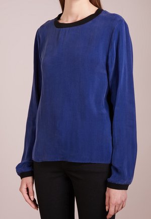 Kobieta ubrana w niebieską bluzkę z długim rękawem, z czarnymi mankietami i kołnierzem, w połączeniu z czarnymi spodniami, stojąca na jednolitym tle.