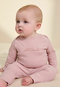 Svetloružový rebrovaný detský outfit s dlhými rukávmi a nohavicami, s vyšitým logom "All Saints" na prednej strane a zaoblenými manžetami.