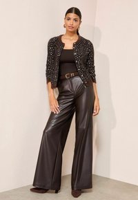 Barna textúrájú kardigán gombokkal, fekete testhez simuló felső, széles lábú barna bőrnadrág és barna magassarkú cipő, világos háttér előtt.