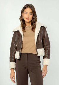 Giacca cropped in pelle marrone con colletto e polsini in shearling, dotata di tasche con zip. Il modello indossa un top beige e pantaloni marroni.