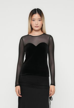 Jongeman met steil zwart en blond ombre haar, gekleed in een zwarte sheer lange mouwen top, laag over een fluwelen bustier.
