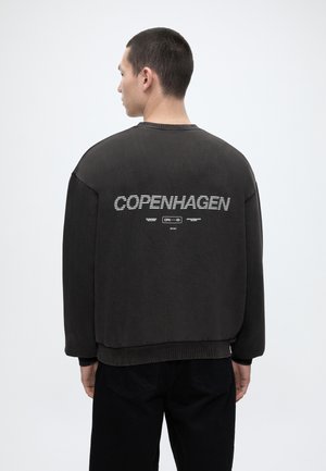 Άνδρας με πλάτη που φοράει μαύρο φούτερ με λευκή επιγραφή "COPENHAGEN" στην πλάτη, σε λευκό φόντο.