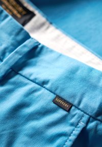 Pantalones cortos de tela azul claro con una textura suave, que cuentan con una etiqueta negra con "SUPERDRY" en letras doradas en la cintura.