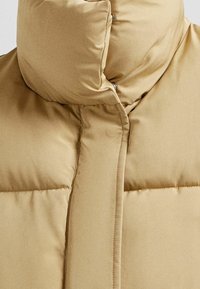 Hnědá péřová bunda s vysokým límcem, lesklou tkaninou, elegantním prošíváním a patentkami na přední straně pro zapnutí.