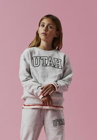 Sivi dukserica i trenirke s natpisom "UTAH", s crvenim i bijelim prugastim detaljima na manžetama i pojasu, meka tekstura. Ružičasta pozadina.