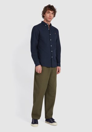 Camicia blu navy con maniche lunghe, abbinata a pantaloni verdi oliva e sneakers blu navy. Design semplice con un piccolo logo sul petto.