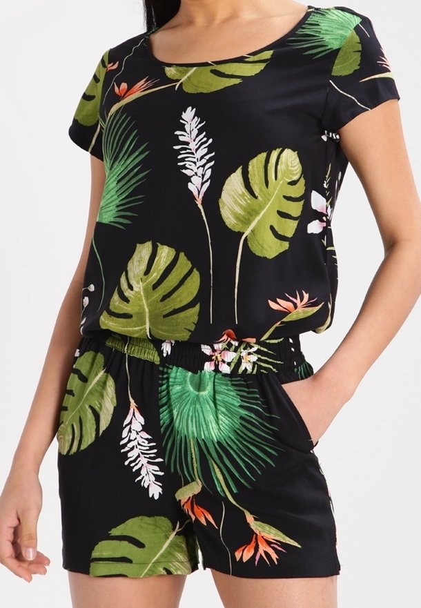 Femme portant une combinaison-short noire à manches courtes avec imprimé de feuilles et fleurs tropicales vertes et orange, une main reposant dans la poche du short.