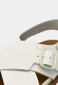 Άσπρο δερμάτινο σανδάλι με σχεδιασμό σταυρωτού λουριού, ρυθμιζόμενο κούμπωμα και καφέ σόλα. Ομαλή υφή με μινιμαλιστική αισθητική.