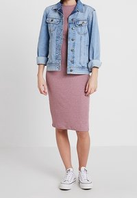 Veste en denim bleu clair sur une robe midi ajustée rose, associée à des baskets blanches. La veste est dotée de boutons et de poches à rabat.