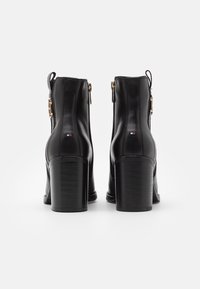 Bottines en cuir noir avec des talons épais, présentant des détails en or et un patch logo décoratif sur le talon, affichant une texture lisse.