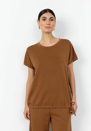 Woman with dark hair wears brown short-sleeve top and matching pants, standing against a plain white background, with gold earrings.