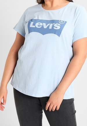 Vrouw met een lichtblauw Levi's T-shirt en een donkere broek, staand met ontspannen armen langs het lichaam tegen een effen achtergrond.