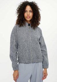 Kobieta z kręconymi włosami, ubrana w szary teksturowany kurtkę z zamkiem i jasnoszare spodnie, stojąca na tle jednolitego białego tła.