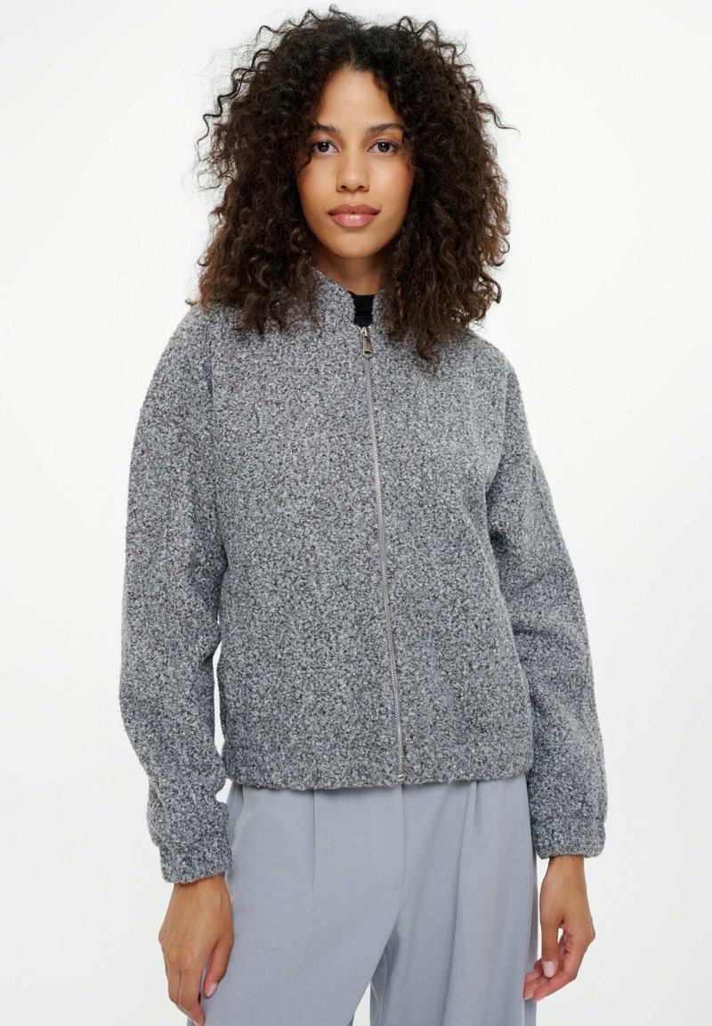 Kobieta z kręconymi włosami, ubrana w szary teksturowany kurtkę z zamkiem i jasnoszare spodnie, stojąca na tle jednolitego białego tła.