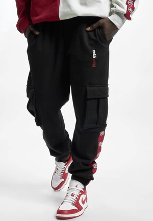 Personne portant un pantalon cargo noir avec une bande latérale rouge, des baskets blanches et rouges, et un sweat-shirt rouge et blanc, mains dans les poches.