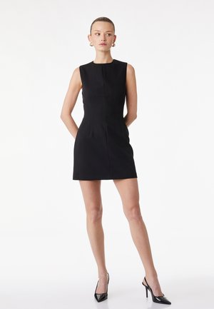 Femme debout, les mains derrière le dos, portant une robe mini noire sans manches et des chaussures slingback à talons hauts noires sur fond blanc.