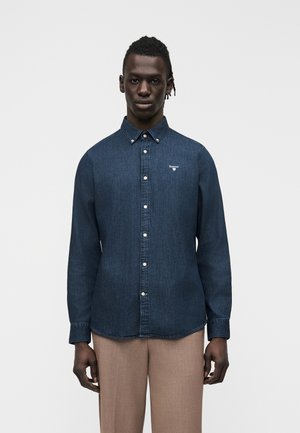 Homme portant une chemise en jean bleu foncé boutonnée avec un petit logo et un pantalon beige, debout devant un fond clair uni.