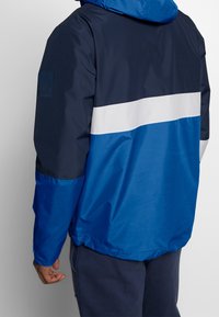Giacca resistente all'acqua con una combinazione di colori blu e navy, dotata di una striscia orizzontale bianca, cappuccio regolabile e polsini elasticizzati.