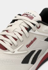 Reebok Nano XV treningssko i lys grå med røde detaljer. Har strukturert overdel, runde lisser og en svart såle. Distinkt logo på siden.