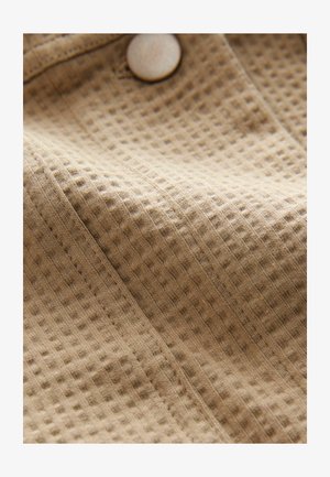 Waffle-patterned beige fabric with visible texture, featuring a light-colored button and stitched seams, showcasing its soft, woven appearance.