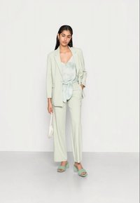 Fato ajustado verde claro com um blazer longo e calças de cintura alta, combinado com um top em tie-dye menta e chinelos verdes de salto. Inclui uma bolsa branca.